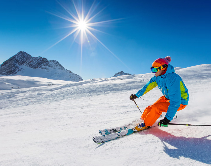 A skier wearing bright clothing navigates a downhill slope on a sunny, clear day, with snowy mountains in the background.