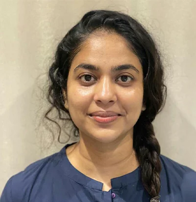 A woman with long, dark hair styled in a loose braid smiles softly at the camera. She is wearing a dark blue blouse and stands in front of a plain, light-colored background.