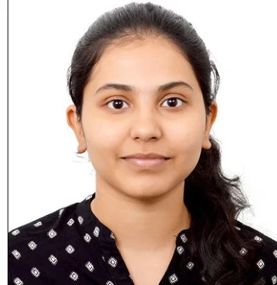 A woman with long dark hair tied back, wearing a black top with white patterns, is looking directly at the camera. She has a neutral expression and a light complexion, set against a plain white background.