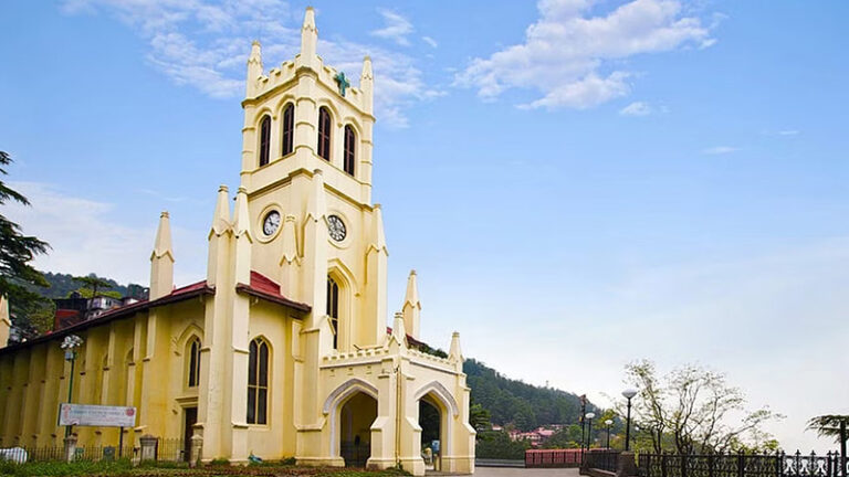 The image shows a historical church with cream-colored walls and a tall clock tower, set against a blue sky with some clouds. The church features Gothic-style architecture, with pointed arches and decorative elements. Surrounding greenery enhances the serene atmosphere.