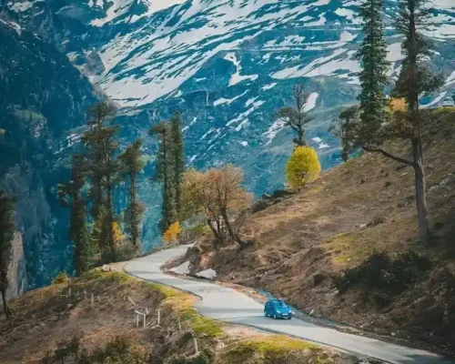 Blue car driving on a winding mountain road with snow-capped peaks